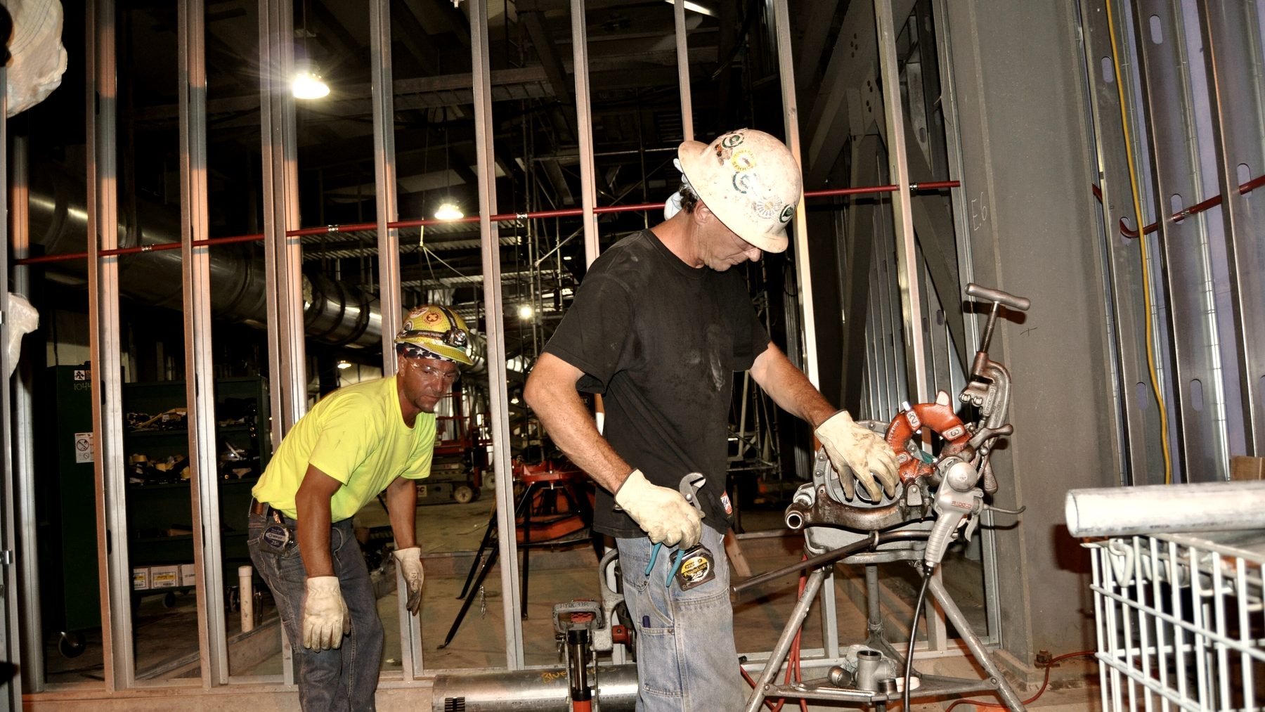 Two tradesmen cutting and threading pipe at a stud-wall rough-in.
