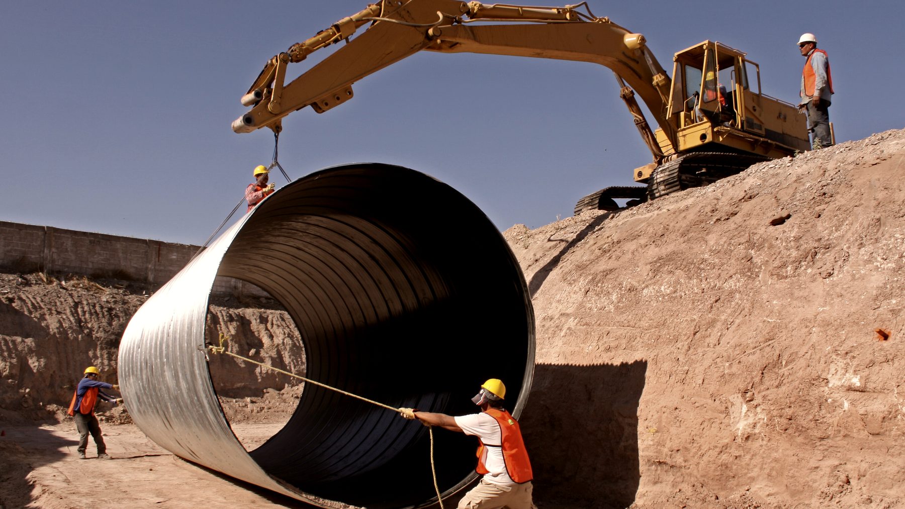 A large pipe being lowered into an open excavation by a tracked excavator.